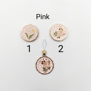 May include: Three round pink enamel charms with gold-tone edges. Item 1 features lily of the valley flowers. Item 2 and the bottom charm display a rose with a bee and small star accents. The bottom charm includes a gold-tone loop and thread attachment.