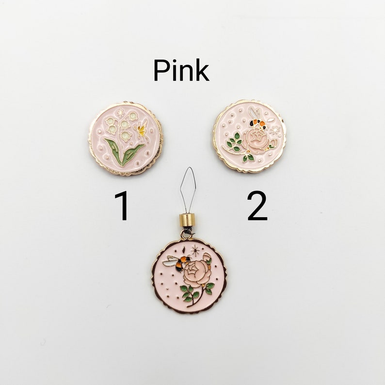May include: Three round pink enamel charms with gold-tone edges. Item 1 features lily of the valley flowers. Item 2 and the bottom charm display a rose with a bee and small star accents. The bottom charm includes a gold-tone loop and thread attachment.