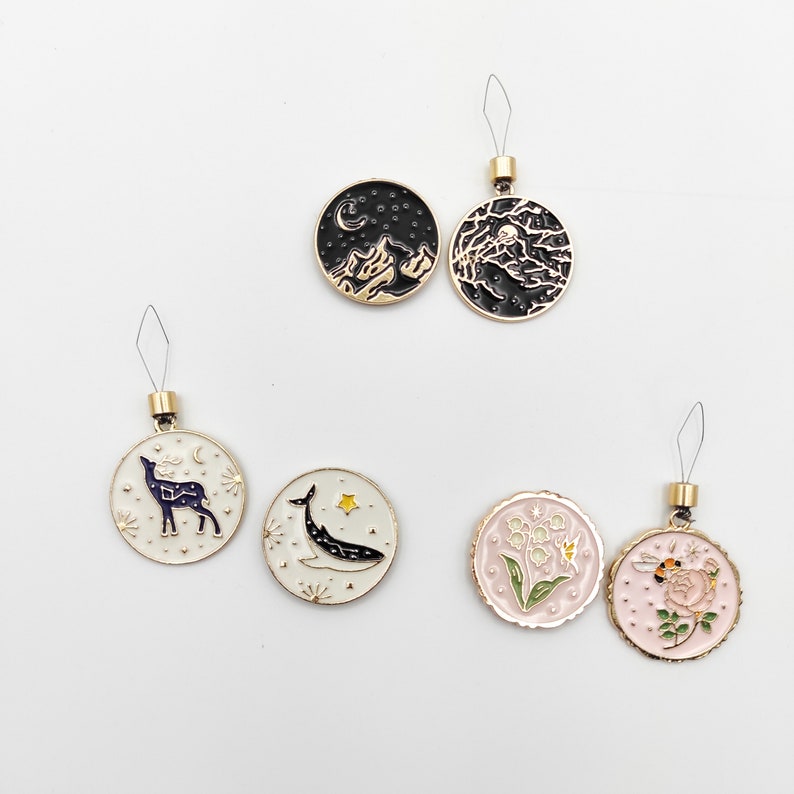 May include: Six circular metal charms with enamel designs. Two black charms feature mountain and moon scenes. Two white charms depict a deer and a whale with stars. Two light pink charms show lily of the valley and a bee on a flower. Each has a gold loop.
