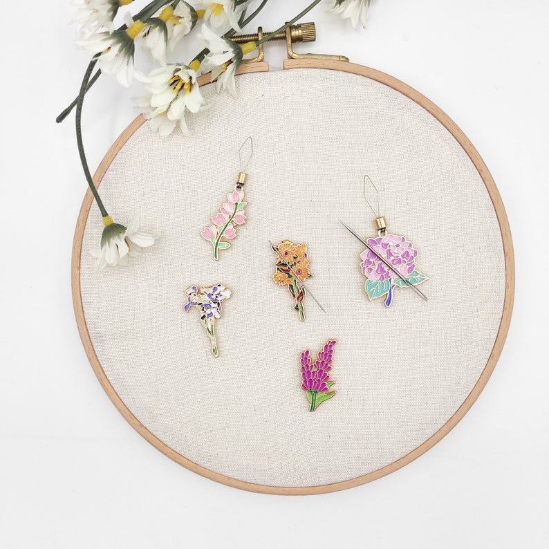 May include: Six colorful flower-shaped needle threaders on a white fabric background. The threaders are made of metal and have a gold finish. Each threader features a different flower design with pink, purple, yellow, and green accents.
