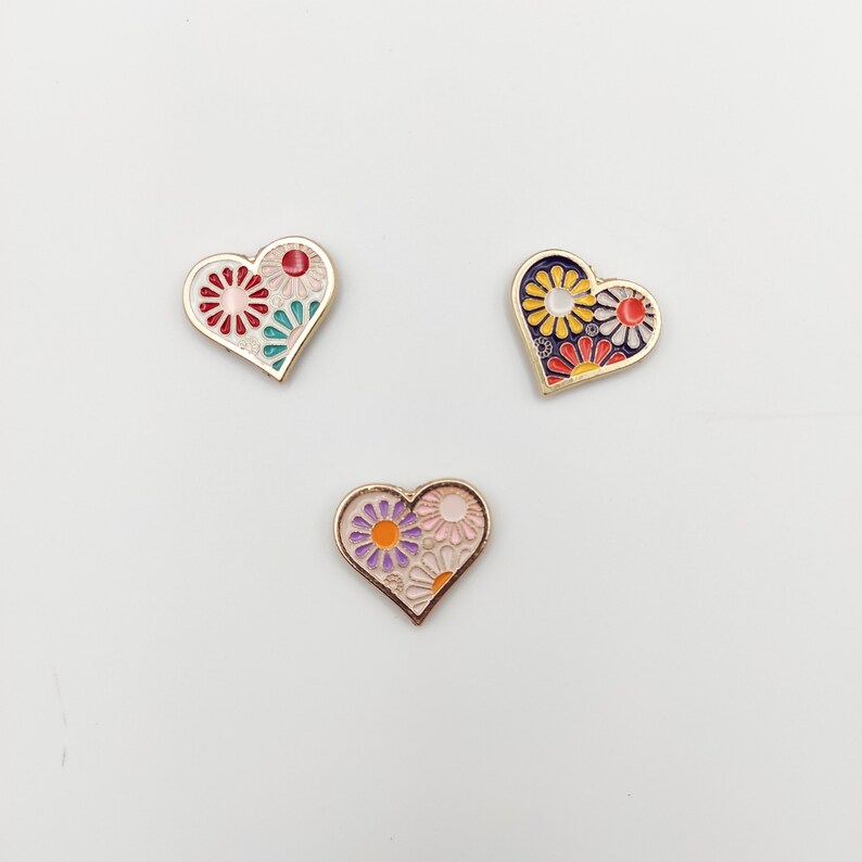 May include: Three gold heart-shaped pins with different floral designs. The first pin has a red, pink, and green floral design. The second pin has a yellow, orange, and red floral design. The third pin has a purple, pink, and orange floral design.