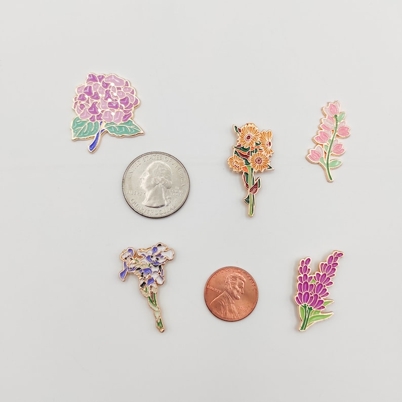 May include: Six enamel pins featuring different floral designs. The pins are arranged in a circle on a white background. The pins are all gold-toned and have colorful enamel details. The flowers include a hydrangea, a bouquet of yellow flowers, a cluster of purple flowers, and two different types of lavender.