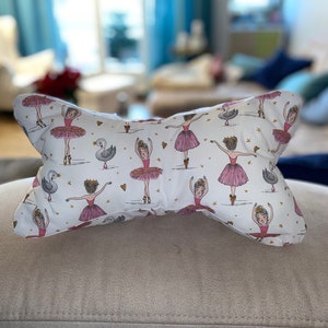 Könnte beinhalten: Ein knochenförmiges Kissen mit weißem Hintergrund, verziert mit Ballerinas in rosa Tutus, Schwänen und goldenen Herzen. Das Kissen liegt auf einer beigen Oberfläche, was auf einen dekorativen oder Komfortartikel hindeutet.