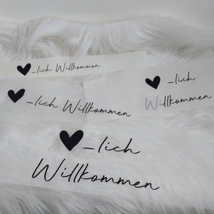 Op de afbeelding: Vier witte stickers met zwarte harten en de tekst "_lich Willkommen" op een wit pluche oppervlak.