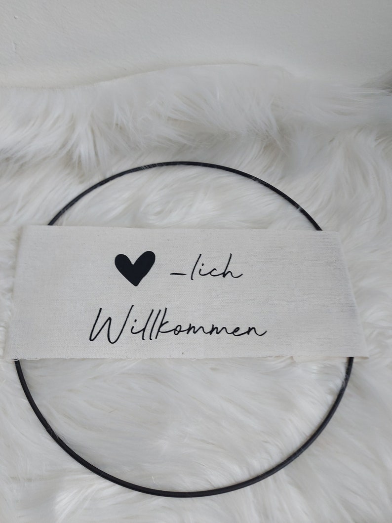 Op de afbeelding: Een wit canvas bord met zwarte tekst die "Herzlich Willkommen" luidt, met een zwart hart voor het woord "Herzlich". Het bord hangt aan een zwarte metalen ring.