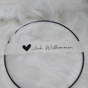Op de afbeelding: Een zwarte metalen ring met een witte stoffen banner waarop "Herzlich Willkommen" staat met een zwart hart.