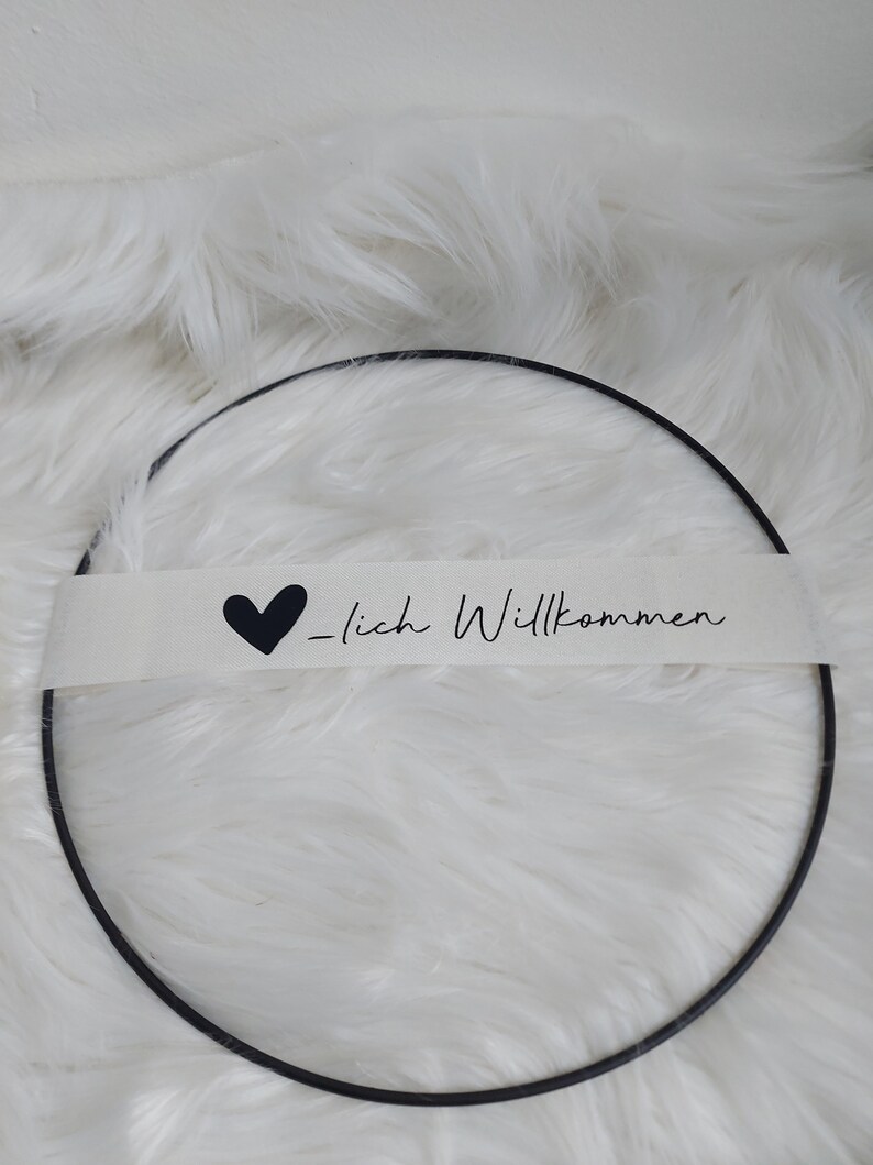 Op de afbeelding: Een zwarte metalen ring met een witte stoffen banner waarop "Herzlich Willkommen" staat met een zwart hart.