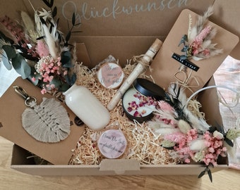 May include: A brown cardboard box filled with gifts for a special occasion. The box is decorated with the words "Herzlichen Glückwunsch" which means "Congratulations" in German. The gifts include a small bouquet of dried flowers, a candle, a small wooden sign, a keychain, and a small jar of bath salts.
