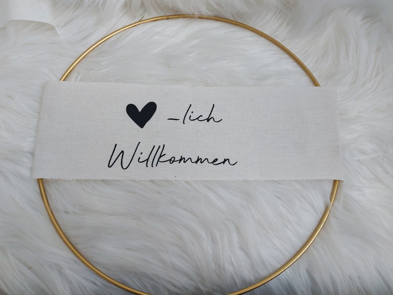 Op de afbeelding: Een gouden hoepel met een witte stoffen banner waarop "Herzlich Willkommen" staat met een zwart hart.