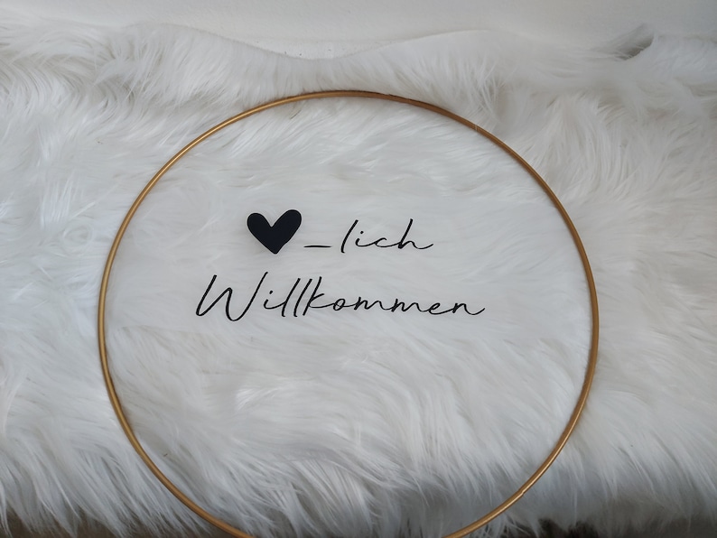 Op de afbeelding: Een gouden hoepel met een zwart hart en de tekst "_lich Willkommen" op een wit pluche oppervlak.