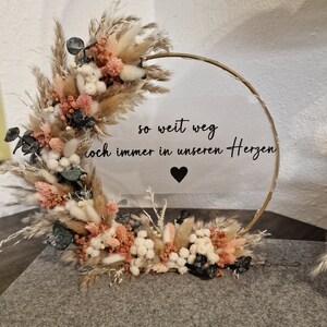 Op de afbeelding: Een gouden hoepel met gedroogde bloemen en groen. Een helder acryl bord met de tekst "so weit weg doch immer in unseren Herzen" en een zwart hart is bevestigd aan de hoepel.
