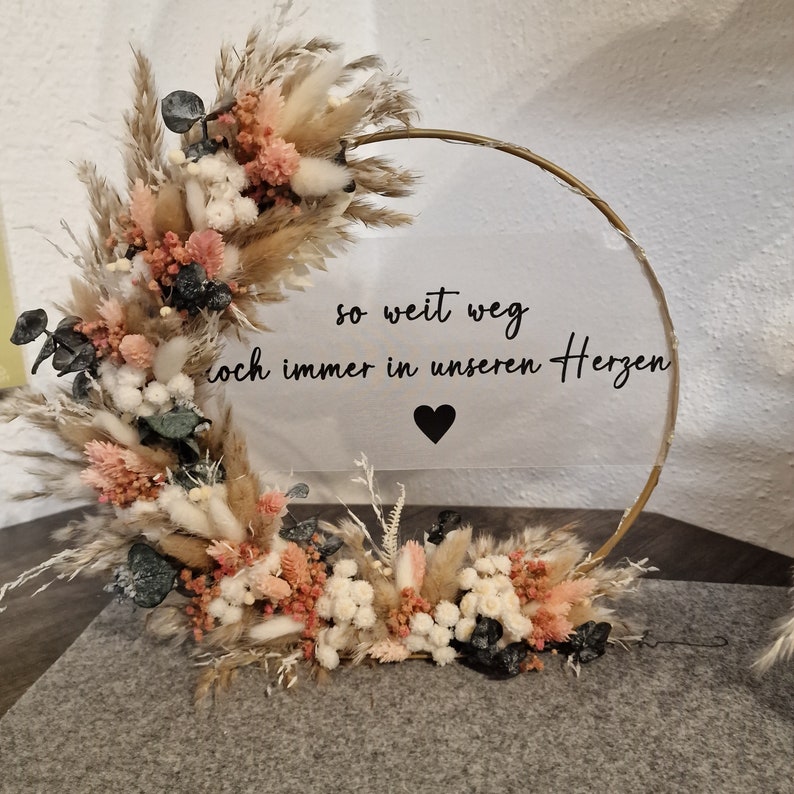Op de afbeelding: Een gouden hoepel met gedroogde bloemen en groen. Een helder acryl bord met de tekst "so weit weg doch immer in unseren Herzen" en een zwart hart is bevestigd aan de hoepel.