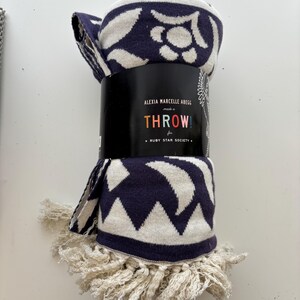 May include: Rolled navy blue and cream throw blanket with a geometric and floral pattern. The throw has a cream fringe and is wrapped with a black label that reads "THROW!" and "ALEXIA MARCELLE ABEGG for RUBY STAR SOCIETY."