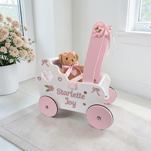 May include: A white and pink wooden toy stroller with the name "Starlette Joy" on the side. The stroller has pink wheels and a pink handle. A teddy bear with a pink bow and flower crown sits inside the stroller.