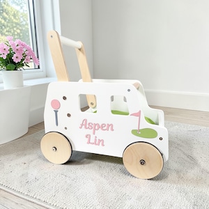 May include: A white wooden golf cart-shaped walker with natural wood wheels and handle. The cart features pink and blue golf-themed graphics and the name "Aspen Lin" in pink lettering. The walker is designed for toddlers.