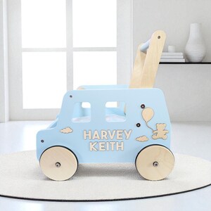 May include: A light blue wooden baby walker shaped like a bus, featuring the name HARVEY KEITH in light wood lettering. The side panel is decorated with a small bear holding a balloon and clouds. It has four natural wood wheels and a handle.