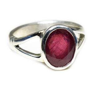 Può includere: Un anello in argento con una grande pietra preziosa rosso scuro di forma ovale, incastonata in una montatura liscia. La fascia dell'anello è divisa in due sezioni distinte che si uniscono ai lati dell'incastonatura centrale.