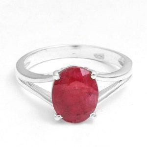 May include: A sterling silver ring featuring a large, oval-shaped, deep red ruby gemstone set in a four-prong mounting. The band is polished silver and splits into two separate shanks as it reaches the central stone, creating a bypass design.