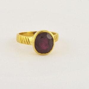 Op de afbeelding: Een gouden ring met een centrale ovaalvormige donkerrode edelsteen in een zetting. De ringband heeft een textuur met een diagonaal geribbeld patroon aan beide zijden van de zetting, wat een contrast vormt met de gladde gouden rand.