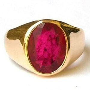 Op de afbeelding: Een gouden ring met een grote, ovaal geslepen, diep roze-rode edelsteen gezet in een gladde, gepolijste gouden band. De steen heeft een gefacetteerd oppervlak dat licht reflecteert en de metalen zetting is dik en solide rondom de steen.