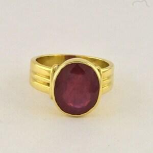 Op de afbeelding: Een gouden ring met een grote, ovaalvormige, donkerrode edelsteen in een gladde zetting. De ringband is van gepolijst goud en heeft aan beide zijden een ontwerp met drie ribbels dat naar de centrale steenzetting toe loopt.