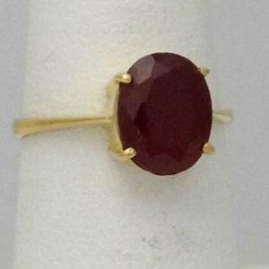 Peut inclure: Une bague en or ornée d'une grande pierre précieuse rouge foncé de taille ovale, fixée par quatre griffes. L'anneau est fin et poli, présentant un design classique qui met en valeur la couleur cramoisie et la surface facettée de la pierre.