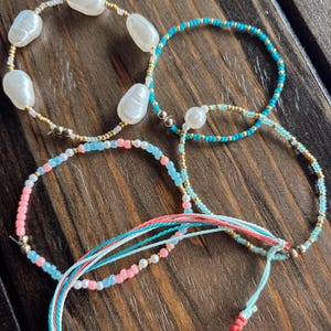May include: A collection of four bracelets featuring white baroque pearls, small gold-tone metallic beads, and turquoise, light blue, and pink glass seed beads. One bracelet includes multiple strands of light blue and pink string with a sliding knot.