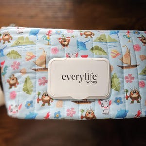 Pode incluir: Uma capa de len&ccedil;os acolchoada azul clara com uma estampa de desenho animado colorida com barcos, flores e personagens. A capa tem uma al&ccedil;a branca e um dispensador de len&ccedil;os de pl&aacute;stico branco com o texto "everylife wipes".