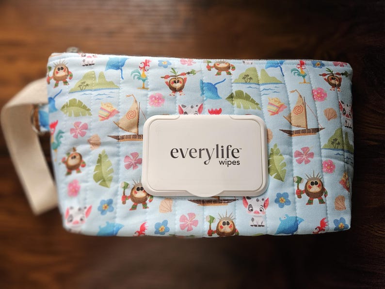 Pode incluir: Uma capa de len&ccedil;os acolchoada azul clara com uma estampa de desenho animado colorida com barcos, flores e personagens. A capa tem uma al&ccedil;a branca e um dispensador de len&ccedil;os de pl&aacute;stico branco com o texto "everylife wipes".