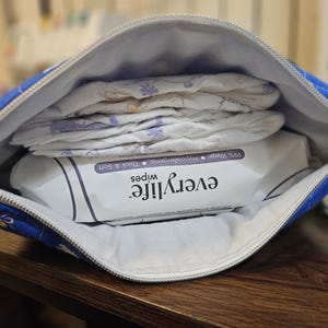 Pode incluir: Uma bolsa com z&iacute;per estampada em azul e branco, aberta para revelar fraldas e um pacote de len&ccedil;os "everylife". Os len&ccedil;os s&atilde;o rotulados como 99% &aacute;gua, hipoalerg&ecirc;nicos, espessos e macios. A bolsa est&aacute; em uma superf&iacute;cie de madeira.