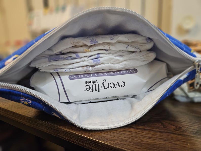 Pode incluir: Uma bolsa com z&iacute;per estampada em azul e branco, aberta para revelar fraldas e um pacote de len&ccedil;os "everylife". Os len&ccedil;os s&atilde;o rotulados como 99% &aacute;gua, hipoalerg&ecirc;nicos, espessos e macios. A bolsa est&aacute; em uma superf&iacute;cie de madeira.