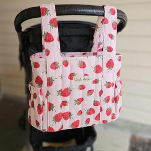 May include: A quilted stroller organizer bag in light pink fabric featuring a repeating pattern of red strawberries with green leaves. The bag has two vertical straps with white snap buttons and a small rectangular label with text attached.