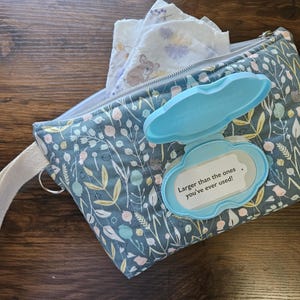 以下が含まれることがあります： 花柄のウェットティッシュケース。水色の蓋と白い持ち手が付いています。ケースは開いており、中にはウェットティッシュが入っています。蓋には「Larger than the ones you've ever used!」と書かれています。