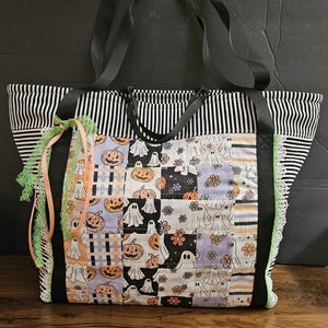 May include: A quilted tote bag featuring a patchwork design of ghosts, pumpkins, and flowers in orange, black, and lavender. The bag has black and white striped side panels, black fabric handles, and decorative green lace and peach ribbon accents.