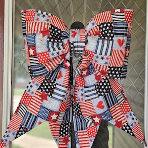May include: A large decorative bow made of fabric featuring a patchwork pattern in red, white, and blue. The design includes various squares with stars, stripes, and heart motifs. The bow has two large loops at the top and two long, pointed tails.