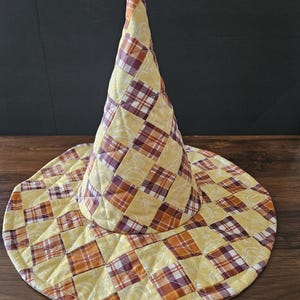 May include: A quilted witch hat featuring a patchwork design of yellow, orange, and maroon plaid squares alternating with light yellow squares decorated with white line drawings of pumpkins and leaves, constructed with a tall cone and wide brim.