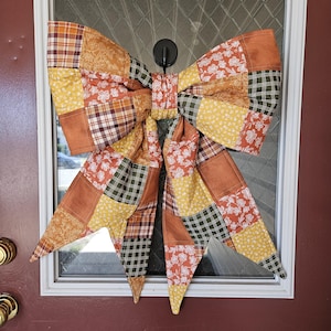 May include: A large decorative bow made from a patchwork of fabric squares in autumn colors including burnt orange, mustard yellow, and forest green. The fabric features various patterns such as plaid, floral prints, and solid textures.