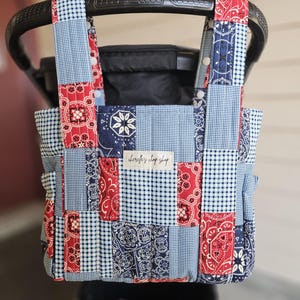 May include: A quilted stroller organizer bag featuring a patchwork design of red, navy blue, and light blue bandana prints and gingham patterns. The bag has two top handles with white snap buttons and a central label reading cherelle's chop shop.