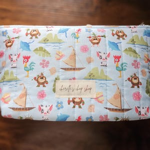 Pode incluir: Uma bolsa de cosm&eacute;ticos acolchoada azul clara com fecho de correr. A bolsa apresenta um padr&atilde;o repetido de personagens de desenhos animados, veleiros e elementos tropicais. Uma etiqueta retangular no centro diz "cherelle's chop shop".