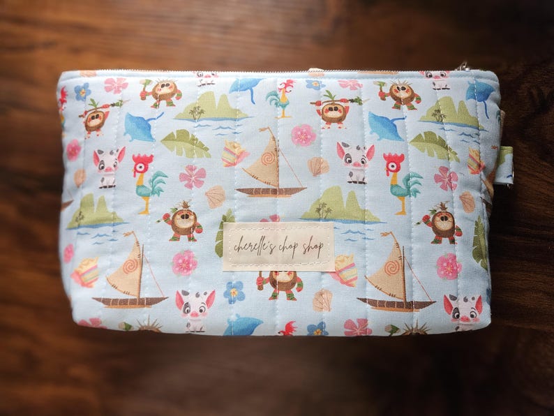 Pode incluir: Uma bolsa de cosm&eacute;ticos acolchoada azul clara com fecho de correr. A bolsa apresenta um padr&atilde;o repetido de personagens de desenhos animados, veleiros e elementos tropicais. Uma etiqueta retangular no centro diz "cherelle's chop shop".