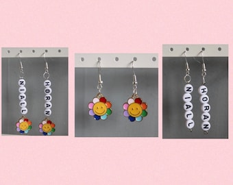 May include: A set of three pairs of earrings. One pair features a rainbow flower with a smiley face, another pair features the name "NIALL" spelled out in white beads, and the last pair features the name "HORAN" spelled out in white beads.