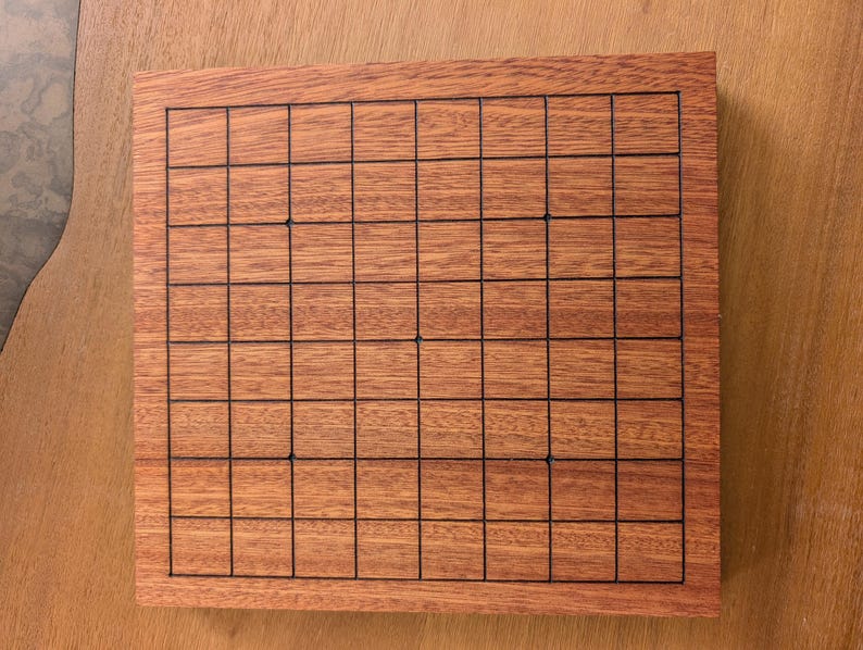 Puede incluir: Un tablero de juego de madera rectangular con una cuadr&iacute;cula de l&iacute;neas negras. El tablero tiene un acabado marr&oacute;n rojizo. La cuadr&iacute;cula est&aacute; formada por l&iacute;neas horizontales y verticales, creando un patr&oacute;n de cuadrados. El tablero es probablemente para un juego de estrategia.