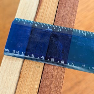 Puede incluir: Una regla azul mide el ancho de tres tiras de madera. Las tiras son de color beige claro, marr&oacute;n medio y marr&oacute;n oscuro. La regla muestra medidas en cent&iacute;metros.