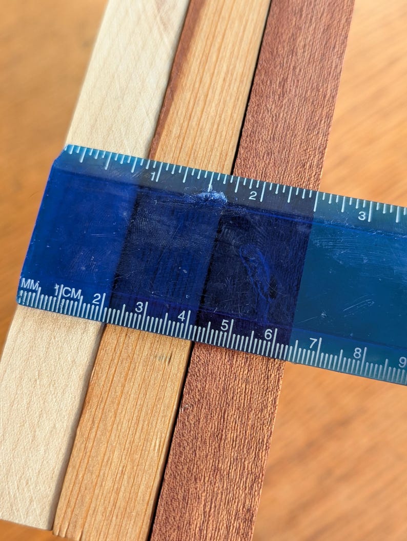 Puede incluir: Una regla azul mide el ancho de tres tiras de madera. Las tiras son de color beige claro, marr&oacute;n medio y marr&oacute;n oscuro. La regla muestra medidas en cent&iacute;metros.