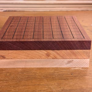 Puede incluir: Un tablero de Go de madera con un patr&oacute;n de cuadr&iacute;cula. El tablero est&aacute; hecho de tres capas de madera en diferentes tonos de marr&oacute;n, con la capa superior con una cuadr&iacute;cula oscura. El tablero es cuadrado.