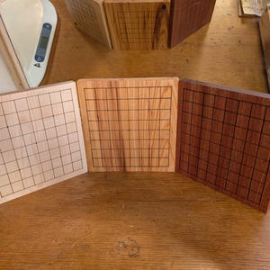Puede incluir: Tres tableros de juego de Go de madera en tonos marrones. Cada tablero presenta una cuadr&iacute;cula de l&iacute;neas negras. Los tableros se exhiben sobre una superficie de madera, mostrando la veta natural. Miden aproximadamente 20 cm.