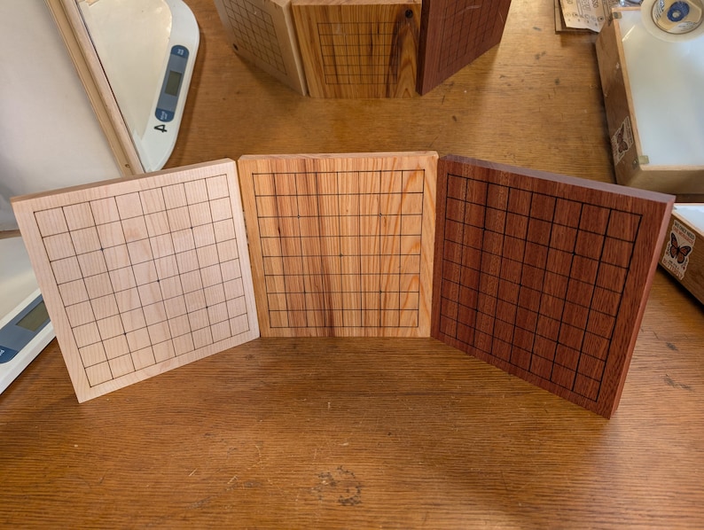 Puede incluir: Tres tableros de juego de Go de madera en tonos marrones. Cada tablero presenta una cuadr&iacute;cula de l&iacute;neas negras. Los tableros se exhiben sobre una superficie de madera, mostrando la veta natural. Miden aproximadamente 20 cm.