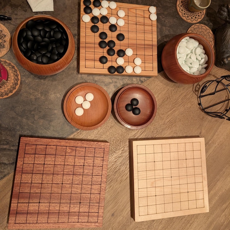 Puede incluir: Un conjunto de juegos de Go, con tableros y cuencos de madera. Los conjuntos incluyen piedras de juego negras y blancas. Los tableros son de madera clara y oscura, con l&iacute;neas de cuadr&iacute;cula. Los cuencos son de madera.