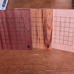 Puede incluir: Tres tableros de juego de madera con un patr&oacute;n de cuadr&iacute;cula. Los tableros est&aacute;n hechos de diferentes tipos de madera, incluyendo un marr&oacute;n rojizo oscuro, un marr&oacute;n claro con vetas y un beige claro. Cada tablero tiene un peque&ntilde;o emblema circular.