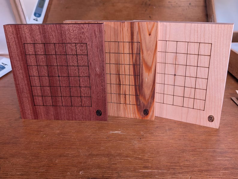 Puede incluir: Tres tableros de juego de madera con un patr&oacute;n de cuadr&iacute;cula. Los tableros est&aacute;n hechos de diferentes tipos de madera, incluyendo un marr&oacute;n rojizo oscuro, un marr&oacute;n claro con vetas y un beige claro. Cada tablero tiene un peque&ntilde;o emblema circular.